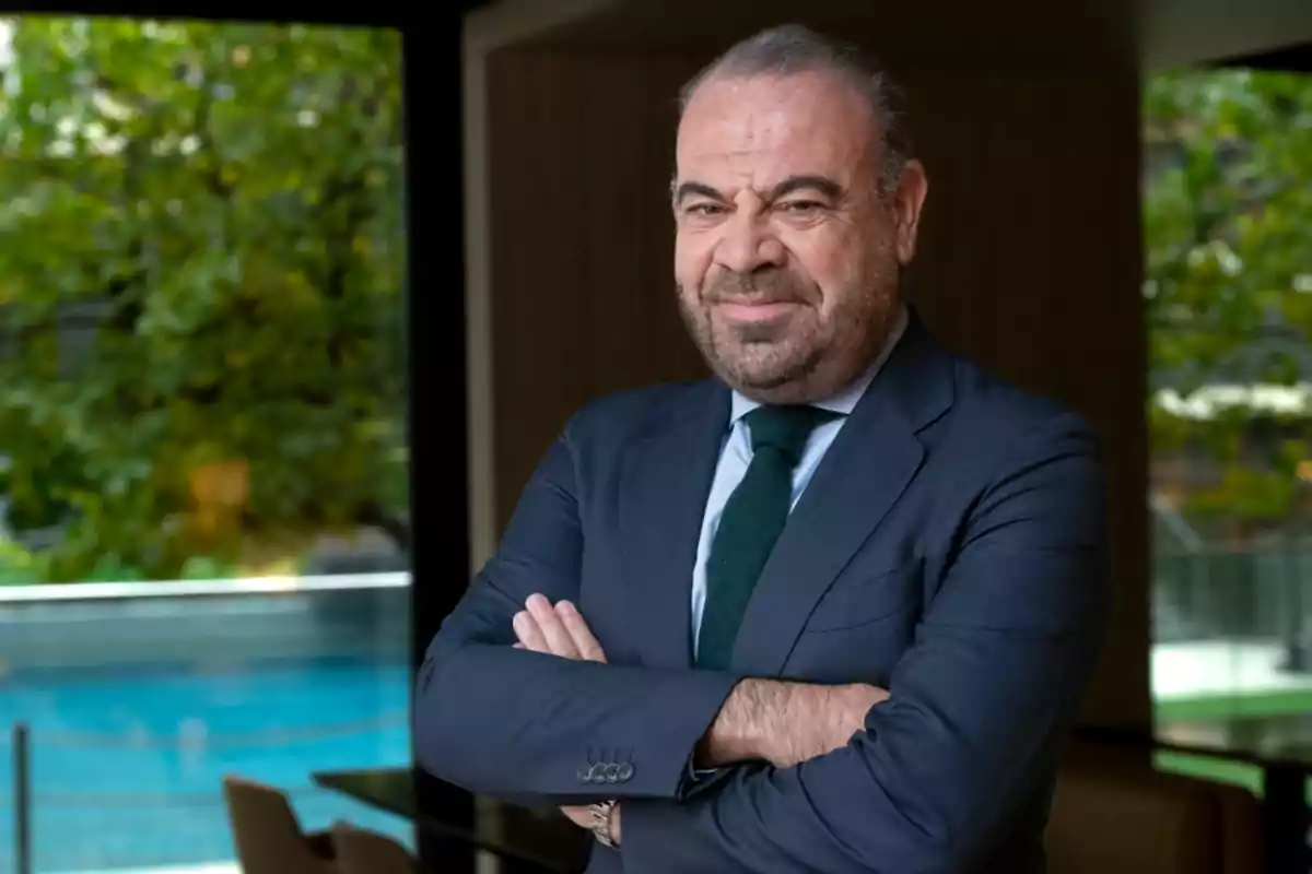 Hombre de traje con los brazos cruzados frente a una ventana con vista a un jardín.