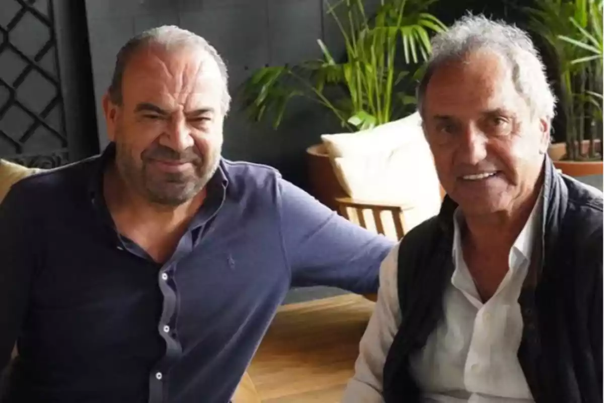 Dos hombres sentados juntos en un ambiente interior con plantas al fondo.