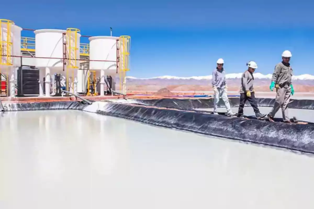 Tres personas con cascos y equipo de seguridad caminan junto a una piscina de evaporación en una planta industrial en un paisaje desértico con montañas al fondo.