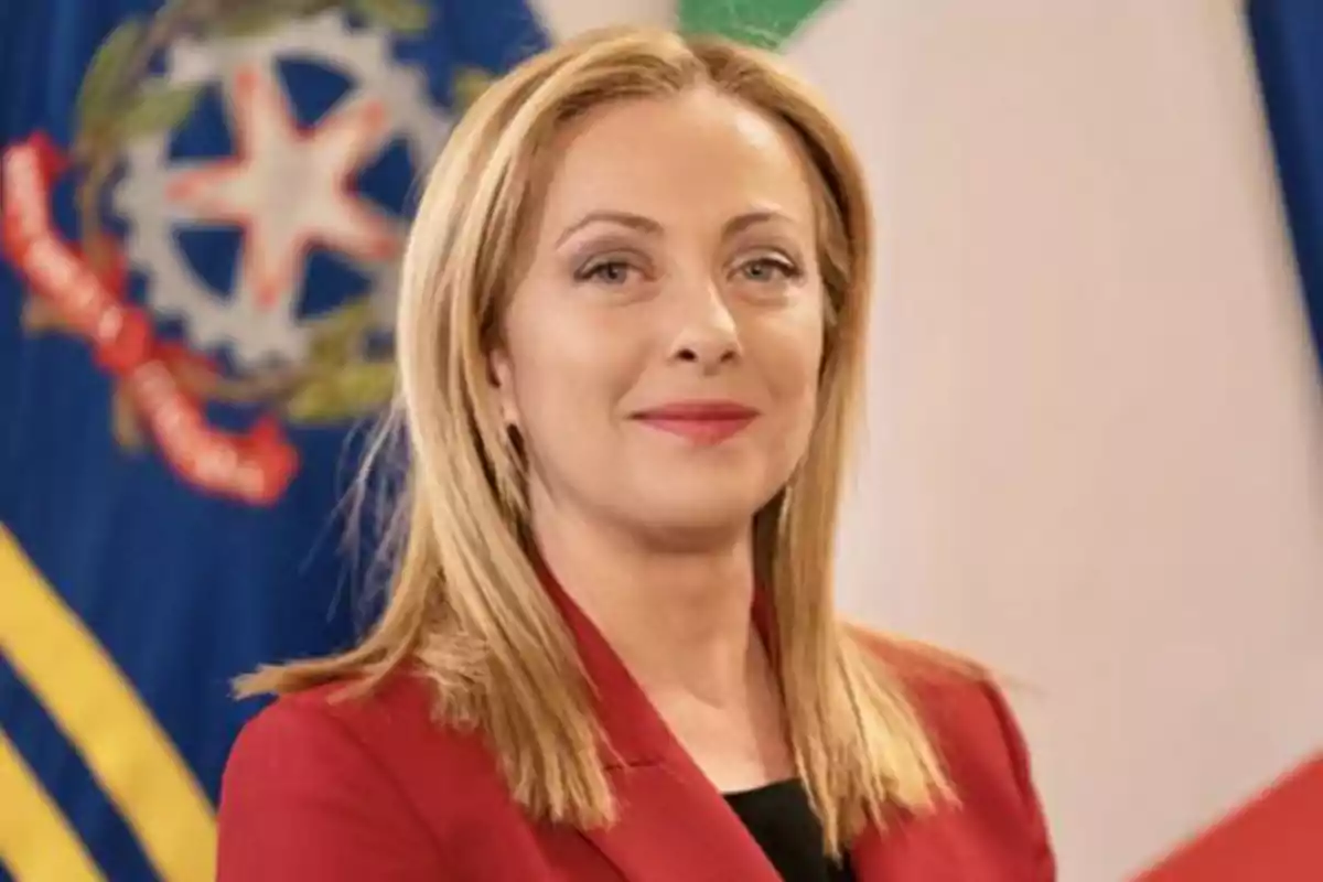 Una persona con cabello rubio y chaqueta roja frente a una bandera.