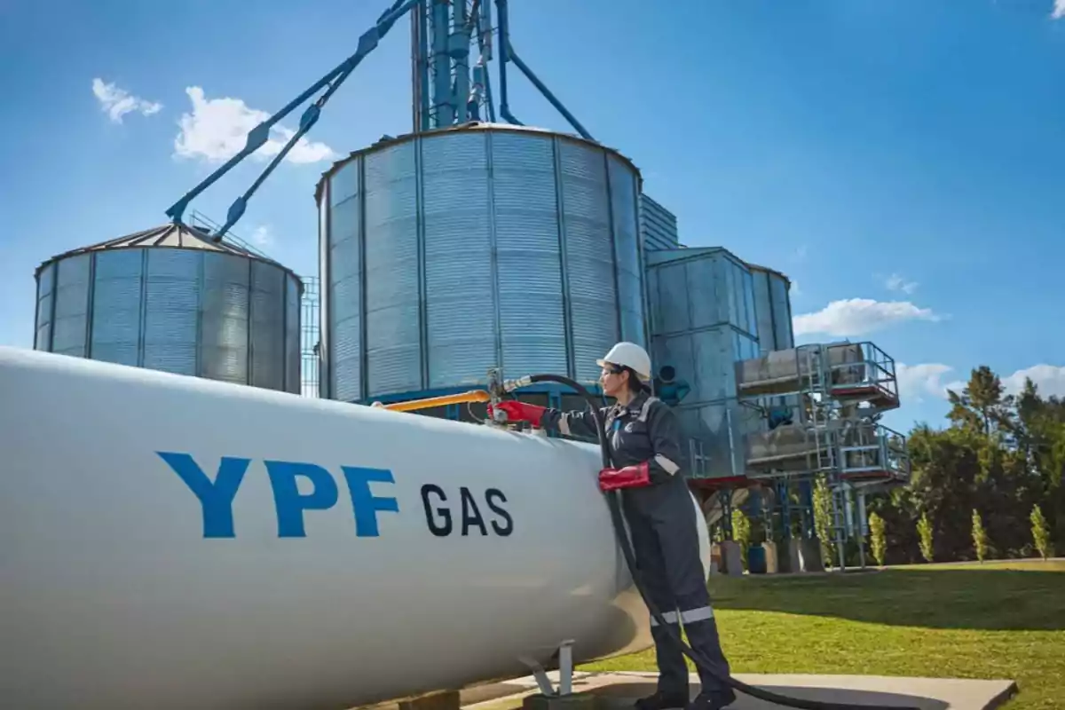 A person with a helmet and red gloves works on a YPF gas tank in front of metal silos under a blue sky.