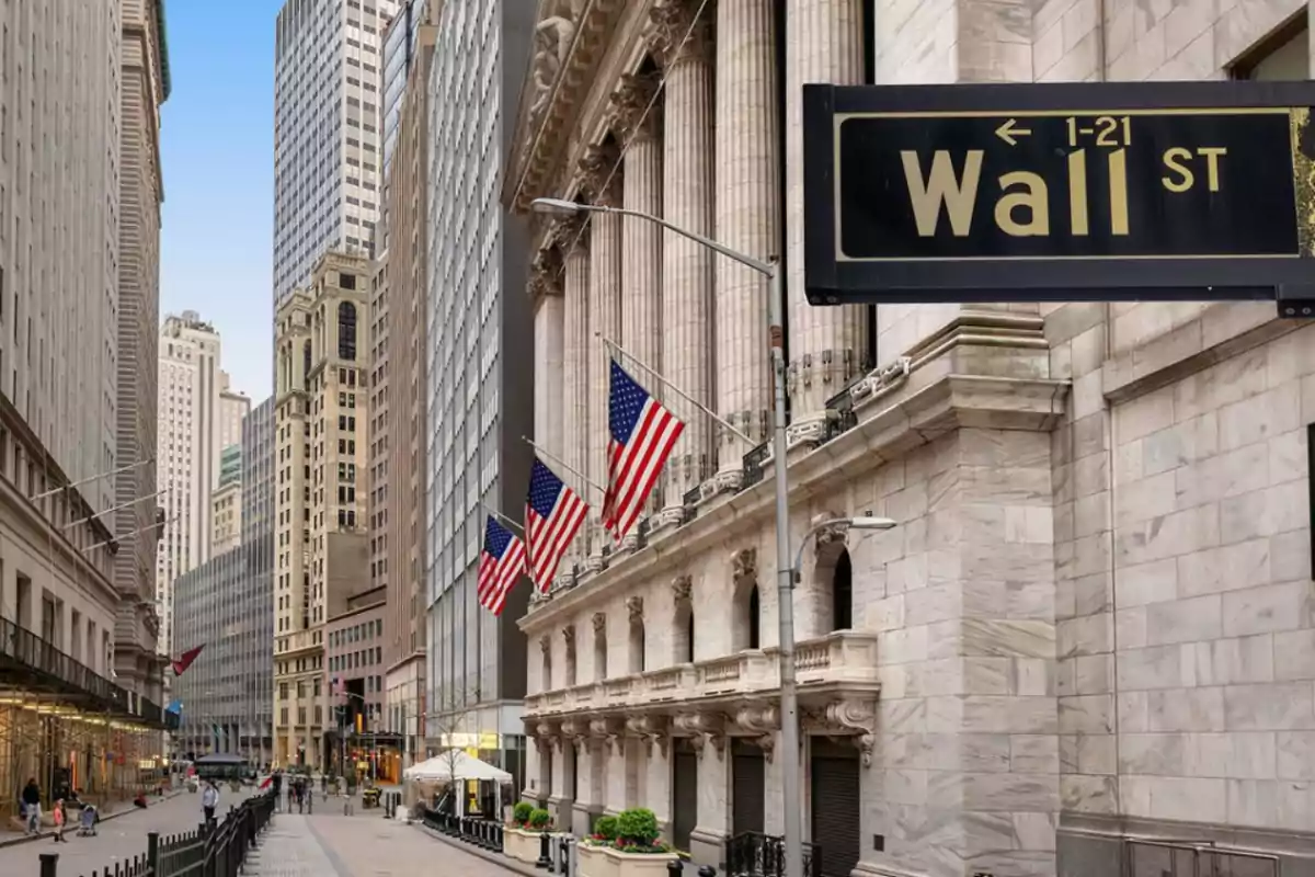 Una calle con edificios altos y banderas estadounidenses, con un letrero que indica Wall Street.