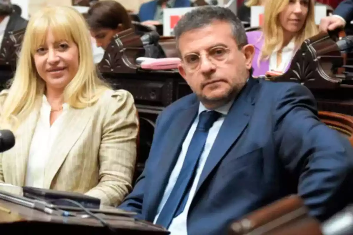 Dos personas sentadas en un entorno formal, posiblemente en una reunión o conferencia, con otras personas al fondo.