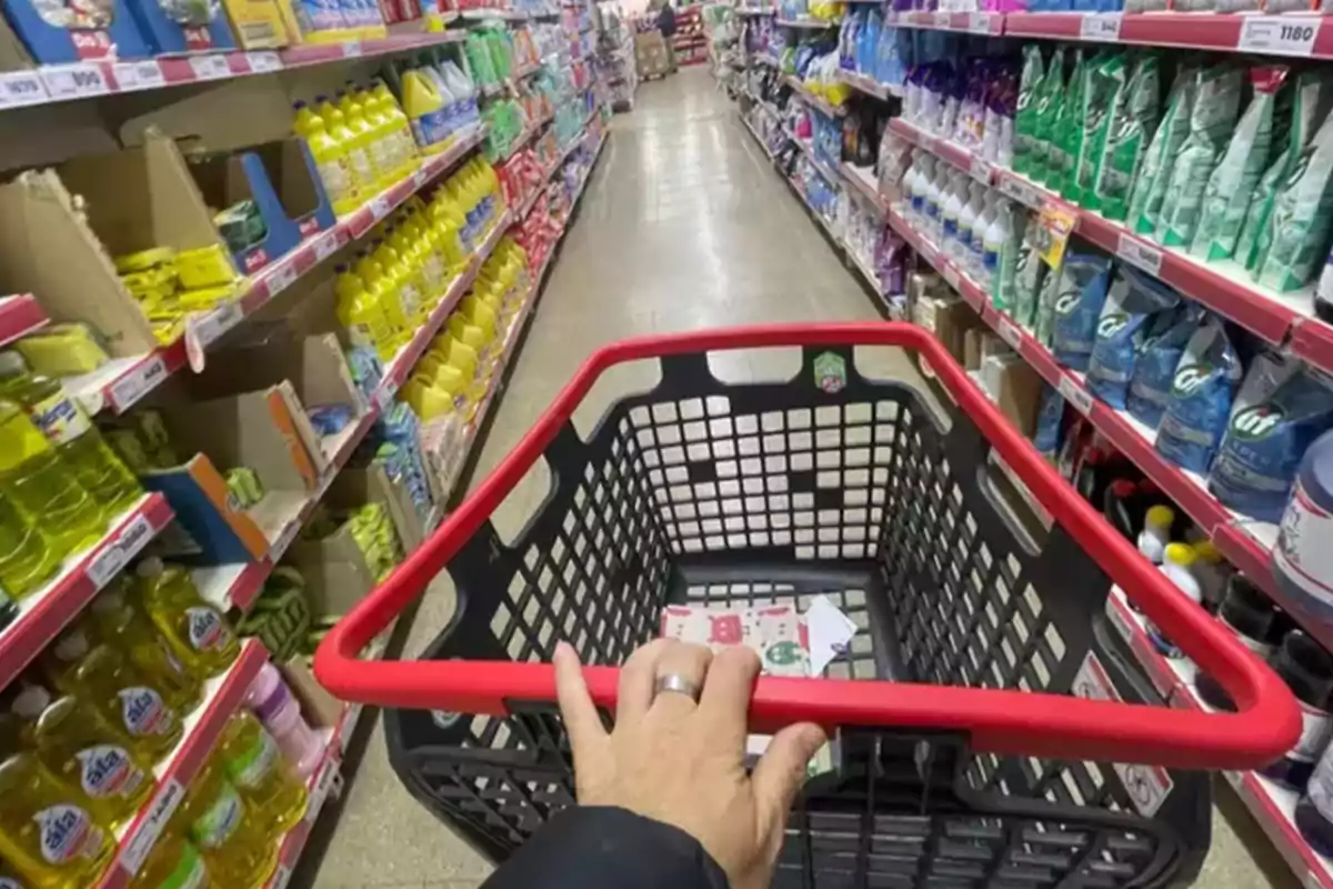 Una persona empuja un carrito de compras por el pasillo de un supermercado lleno de productos de limpieza y artículos para el hogar.