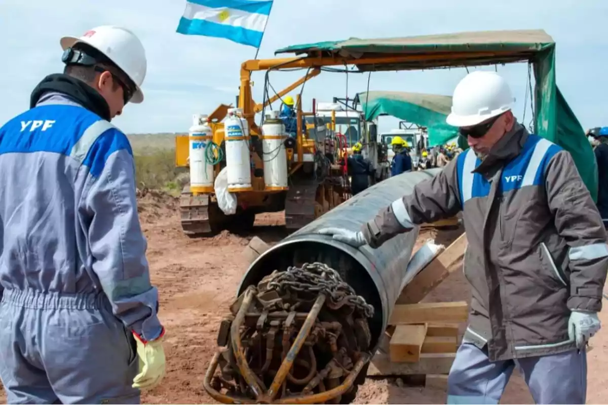 Workers in YPF uniforms inspect a large pipe at a construction site, with machinery and an Argentine flag in the background.