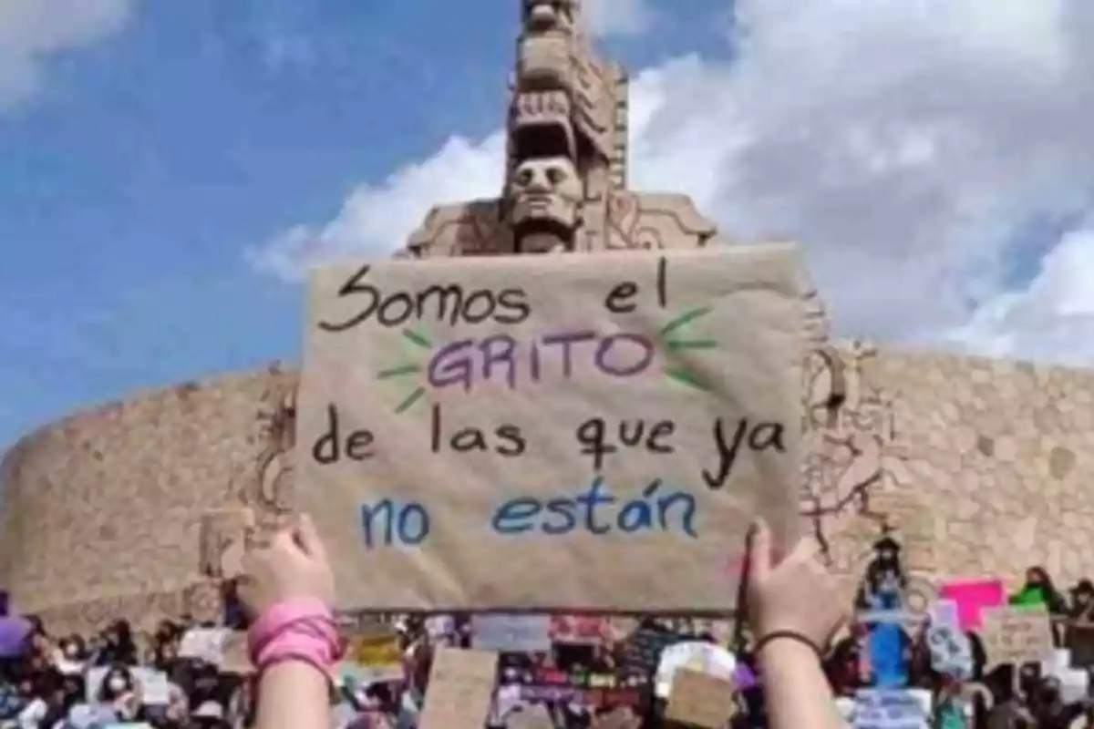 Una multitud sostiene carteles en una manifestación frente a un monumento de piedra, destacando uno que dice "Somos el grito de las que ya no están".