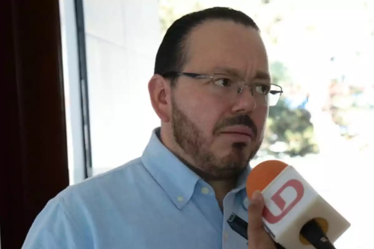 Un hombre con gafas y barba está siendo entrevistado, sosteniendo un micrófono con un logotipo visible.