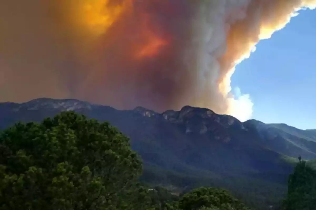 Montañas con un gran incendio forestal en el fondo y humo denso elevándose hacia el cielo.