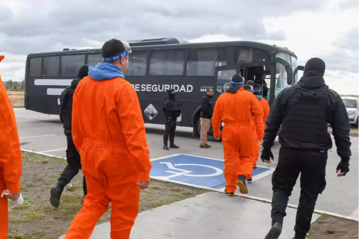 Personas con trajes naranjas y guardias de seguridad se dirigen hacia un autobús en un área de estacionamiento.