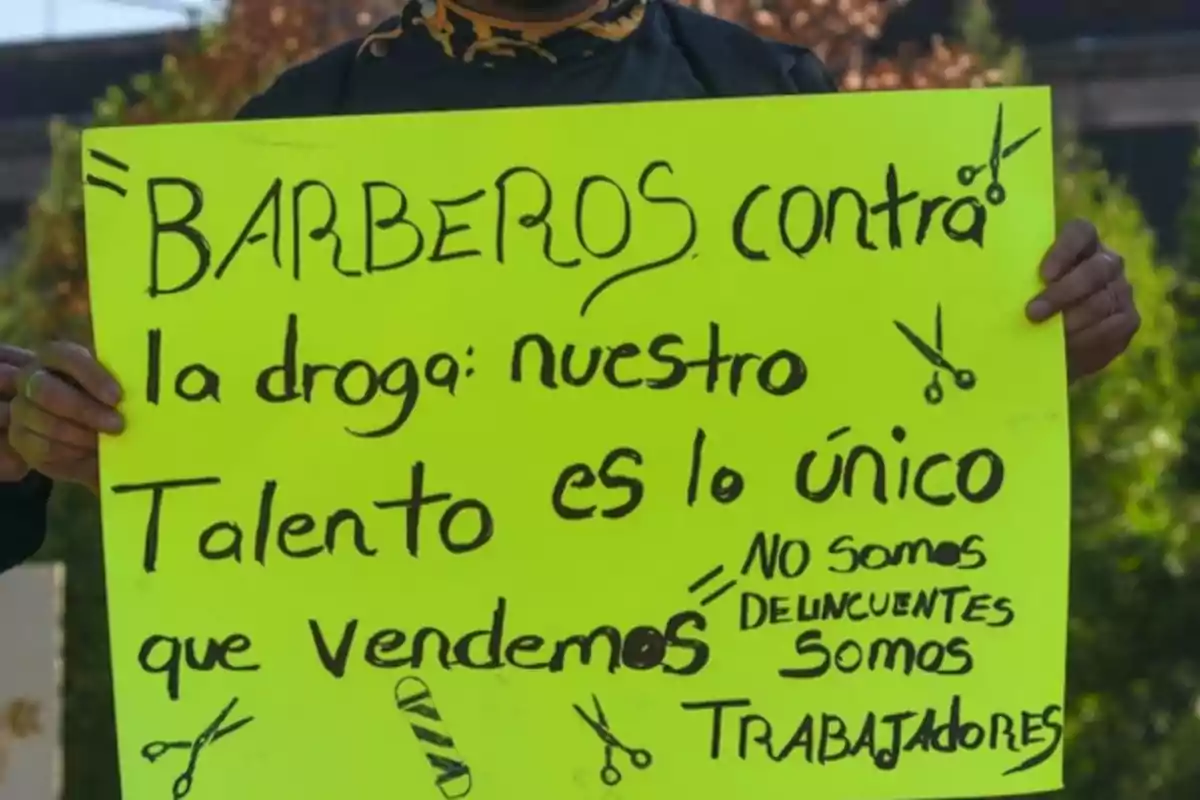 Persona sosteniendo un cartel amarillo que dice "Barberos contra la droga: nuestro talento es lo único que vendemos. No somos delincuentes, somos trabajadores", decorado con dibujos de tijeras y una navaja de barbero.