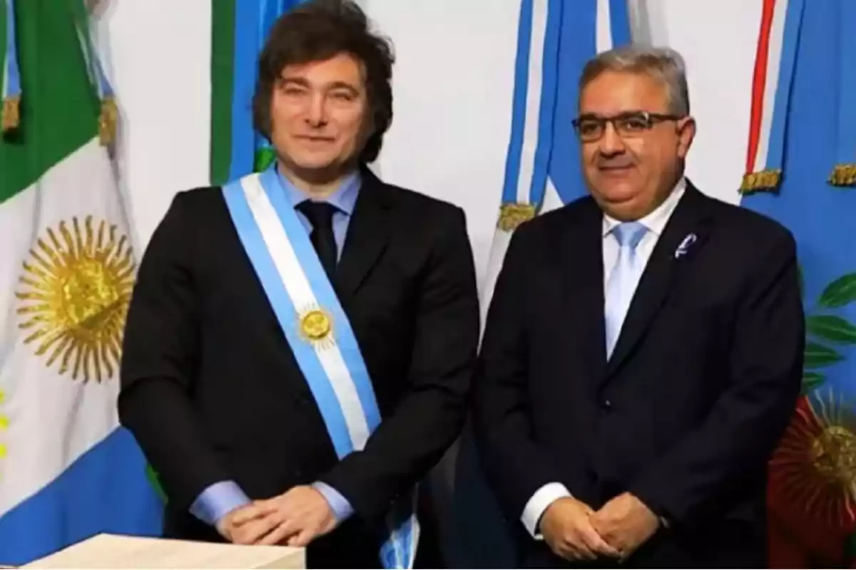 Dos hombres de pie con trajes formales y bandas presidenciales frente a banderas argentinas.