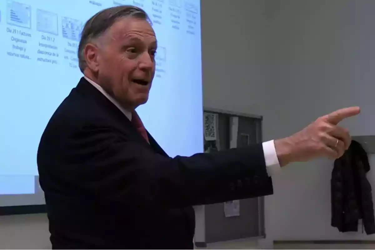 A man in a suit pointing forward in a conference room with a projection screen behind.