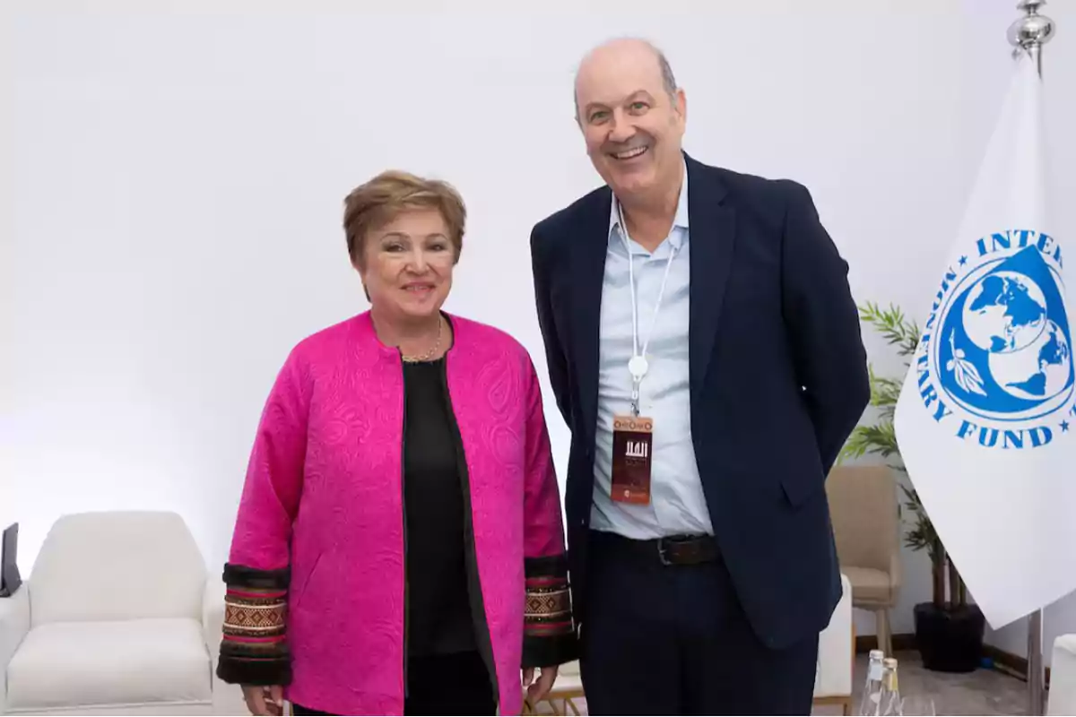 Dos personas posan juntas en un entorno formal con una bandera del Fondo Monetario Internacional al fondo.