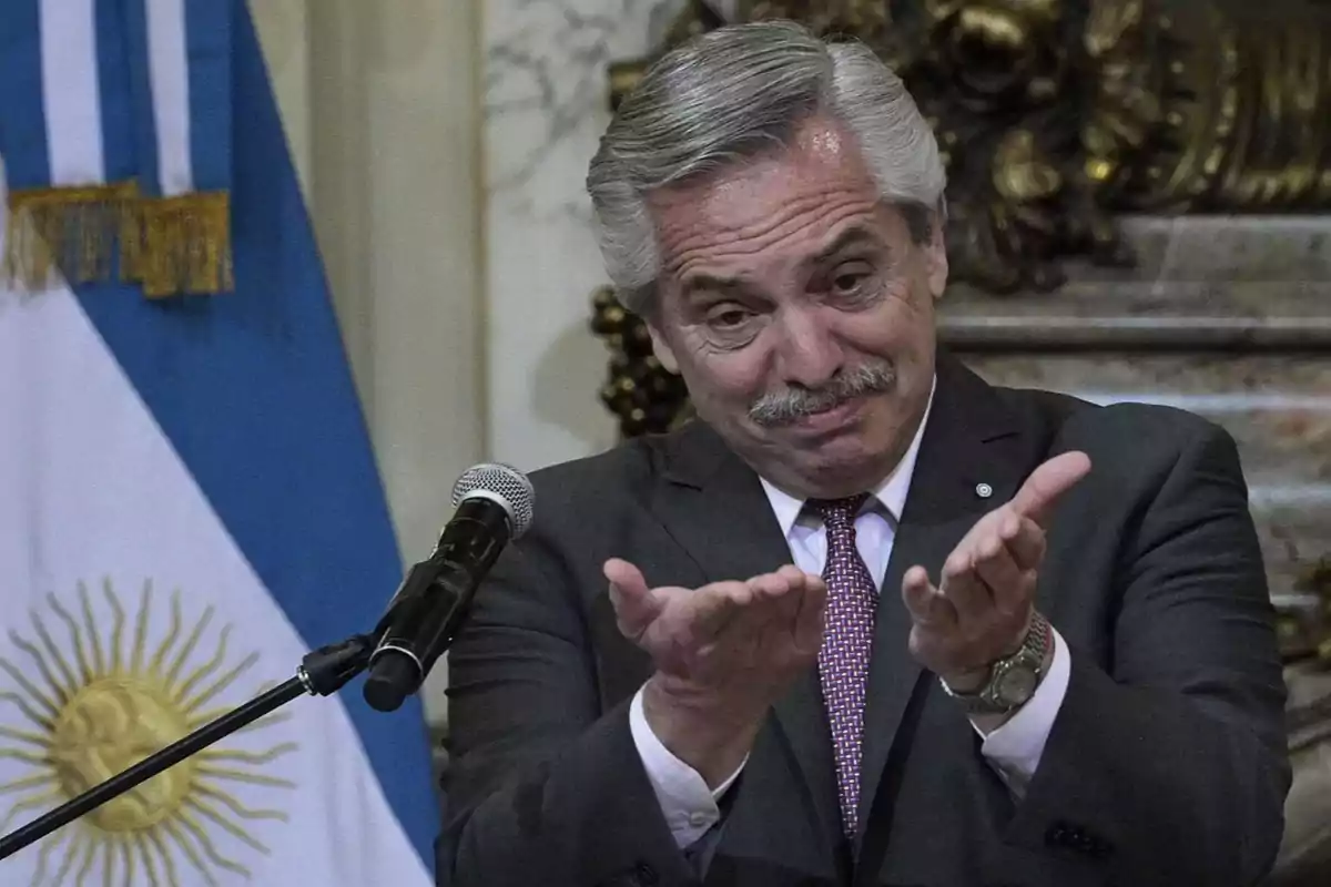 Un hombre de traje gesticula con las manos frente a un micrófono con una bandera de Argentina en el fondo.