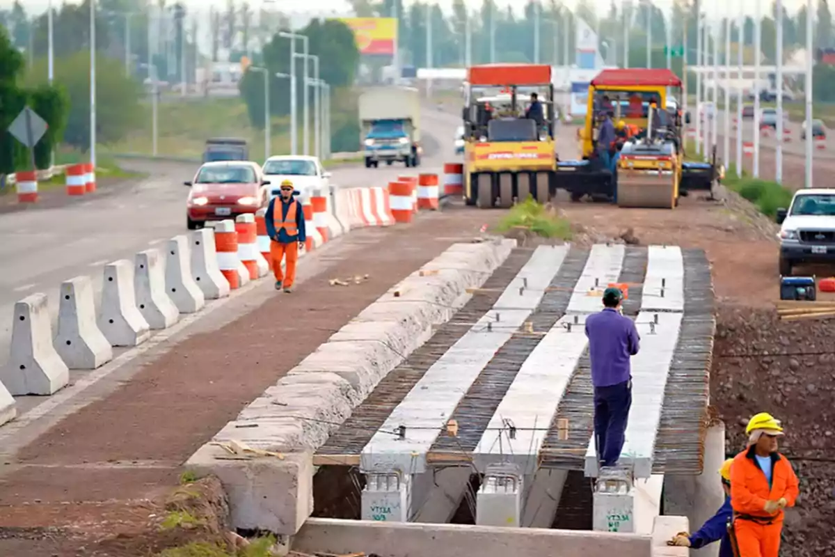 Trabajadores de la construcción en una carretera en reparación, con maquinaria pesada y barreras de seguridad a los lados.