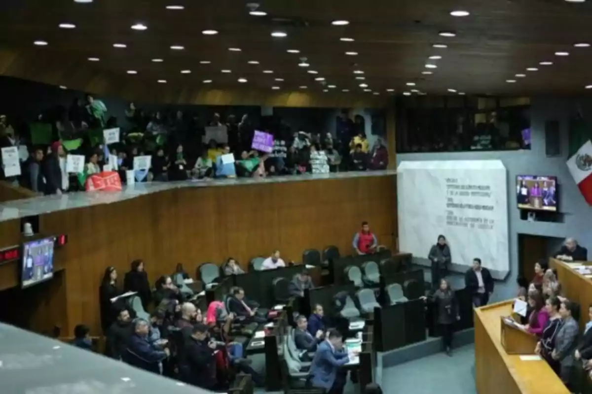 Una sala de asamblea con personas sentadas y de pie, algunas sosteniendo carteles, mientras se lleva a cabo una sesión o reunión formal.