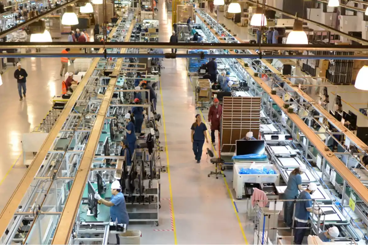 Personas trabajando en una línea de ensamblaje dentro de una fábrica iluminada con luces colgantes.