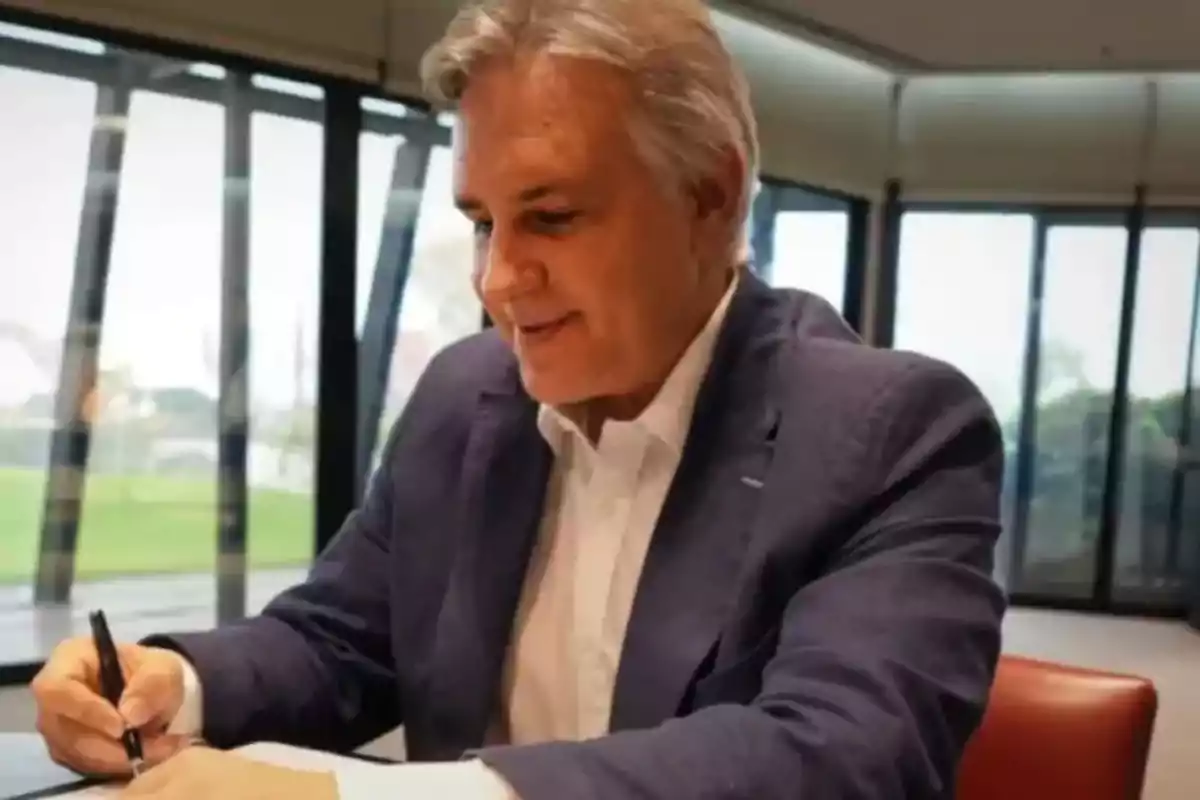 Un hombre de cabello canoso y traje oscuro está escribiendo en un escritorio en una habitación con grandes ventanales.