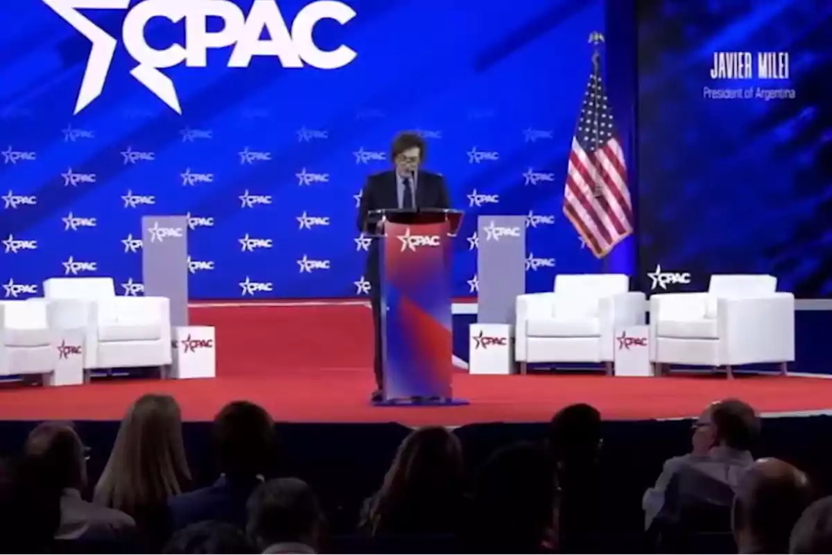 Una persona hablando en un podio en un evento con el logo de CPAC en el fondo y una bandera de Estados Unidos a la derecha.