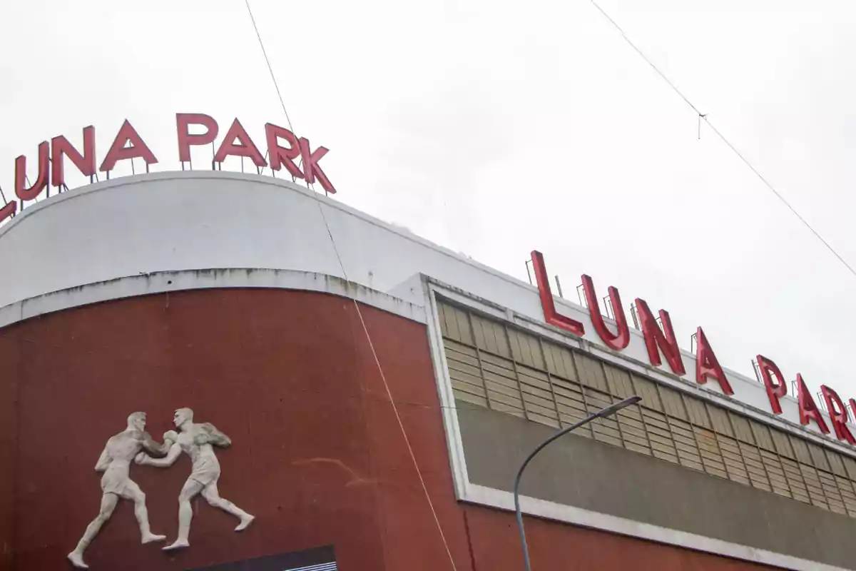 La imagen muestra la fachada de un edificio con el letrero "Luna Park" en letras rojas y una escultura de dos boxeadores en relieve sobre una pared de color marrón.