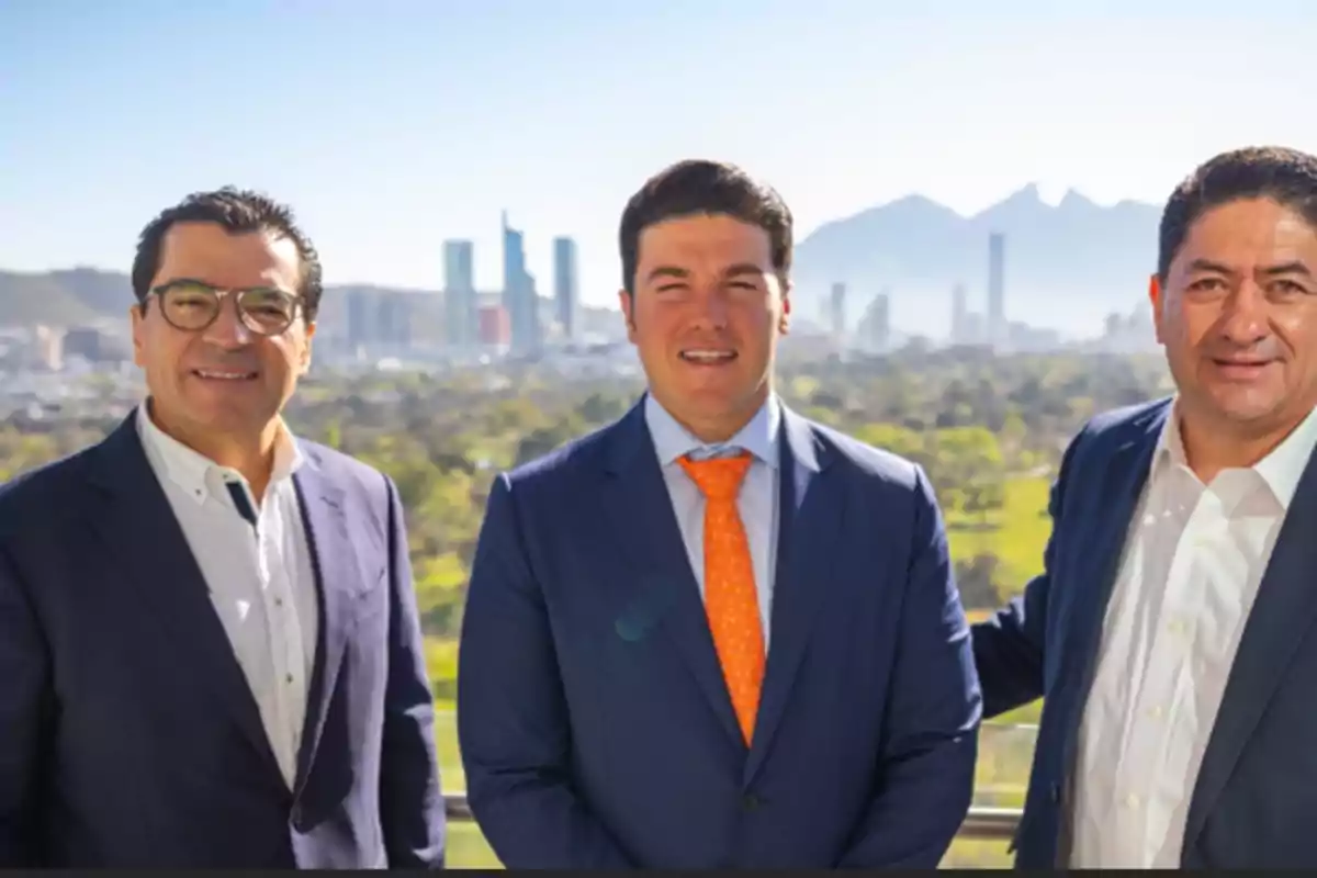 Tres hombres con trajes formales posan al aire libre con un paisaje urbano y montañas al fondo.