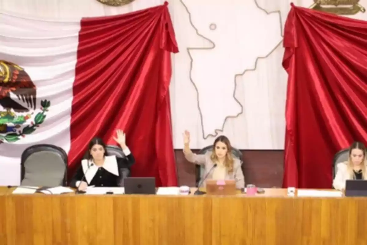 Tres personas sentadas en una mesa levantan la mano en un salón con una bandera de México de fondo.