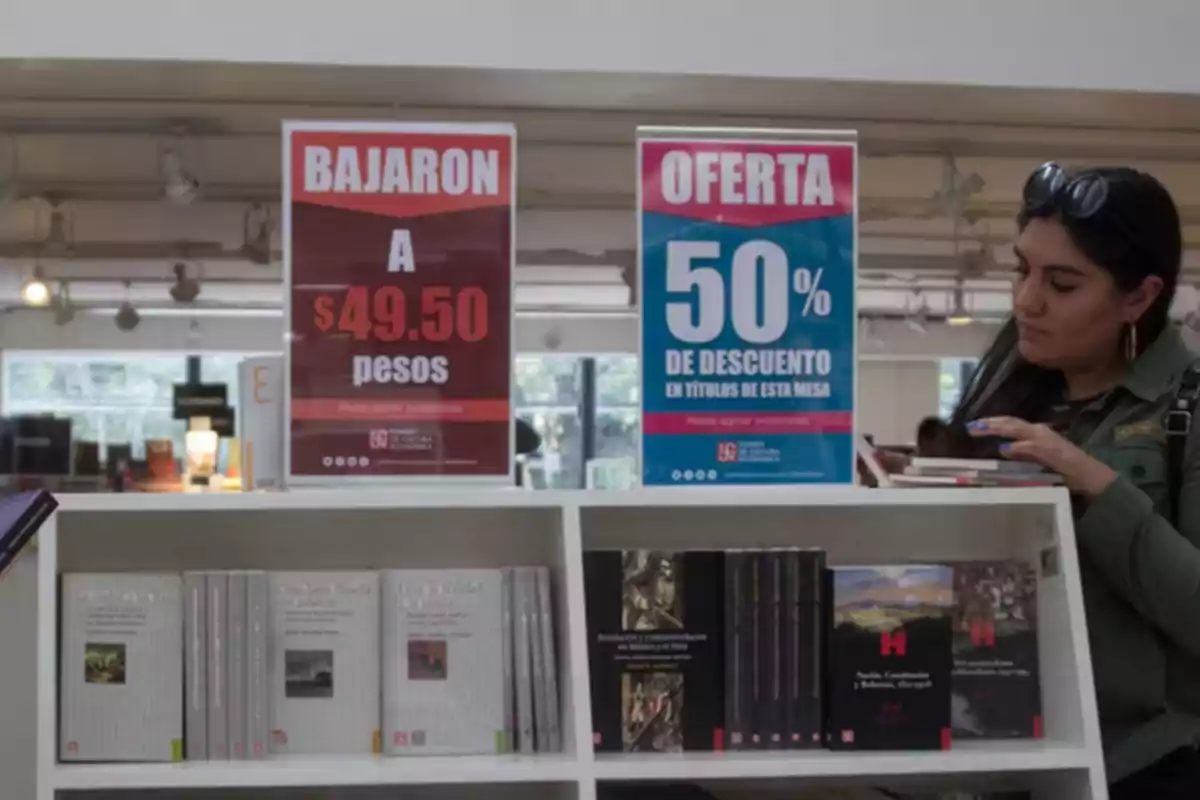 Una mujer revisa libros en una librería que tiene carteles de ofertas, uno indica que los precios bajaron a $49.50 pesos y otro ofrece un 50% de descuento en títulos de una mesa.