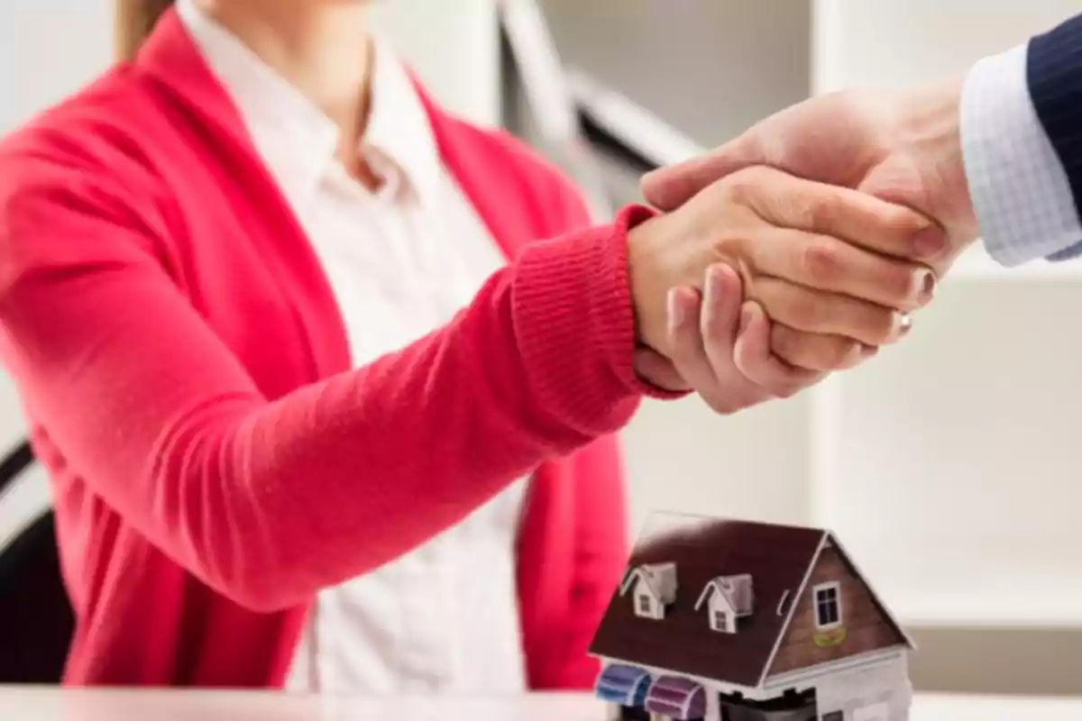 Dos personas estrechándose la mano sobre una mesa con una maqueta de casa.