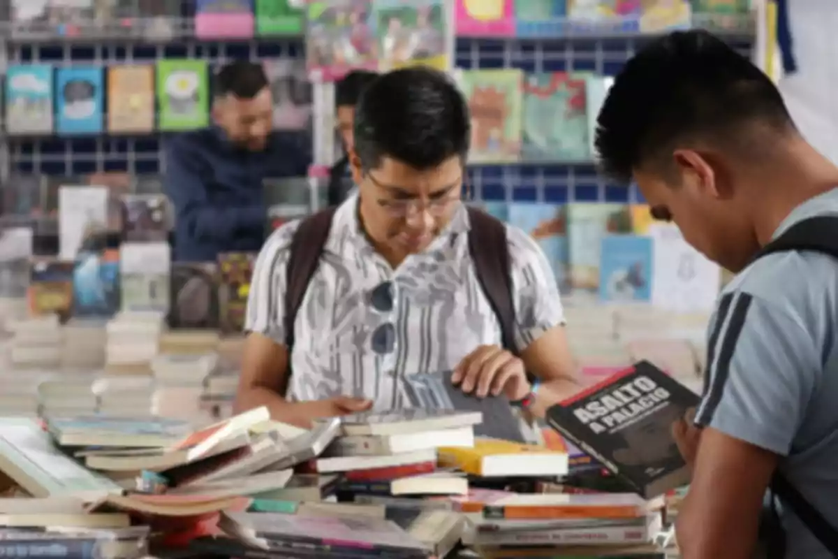 Dos personas revisan libros en una librería con estantes llenos de libros al fondo.