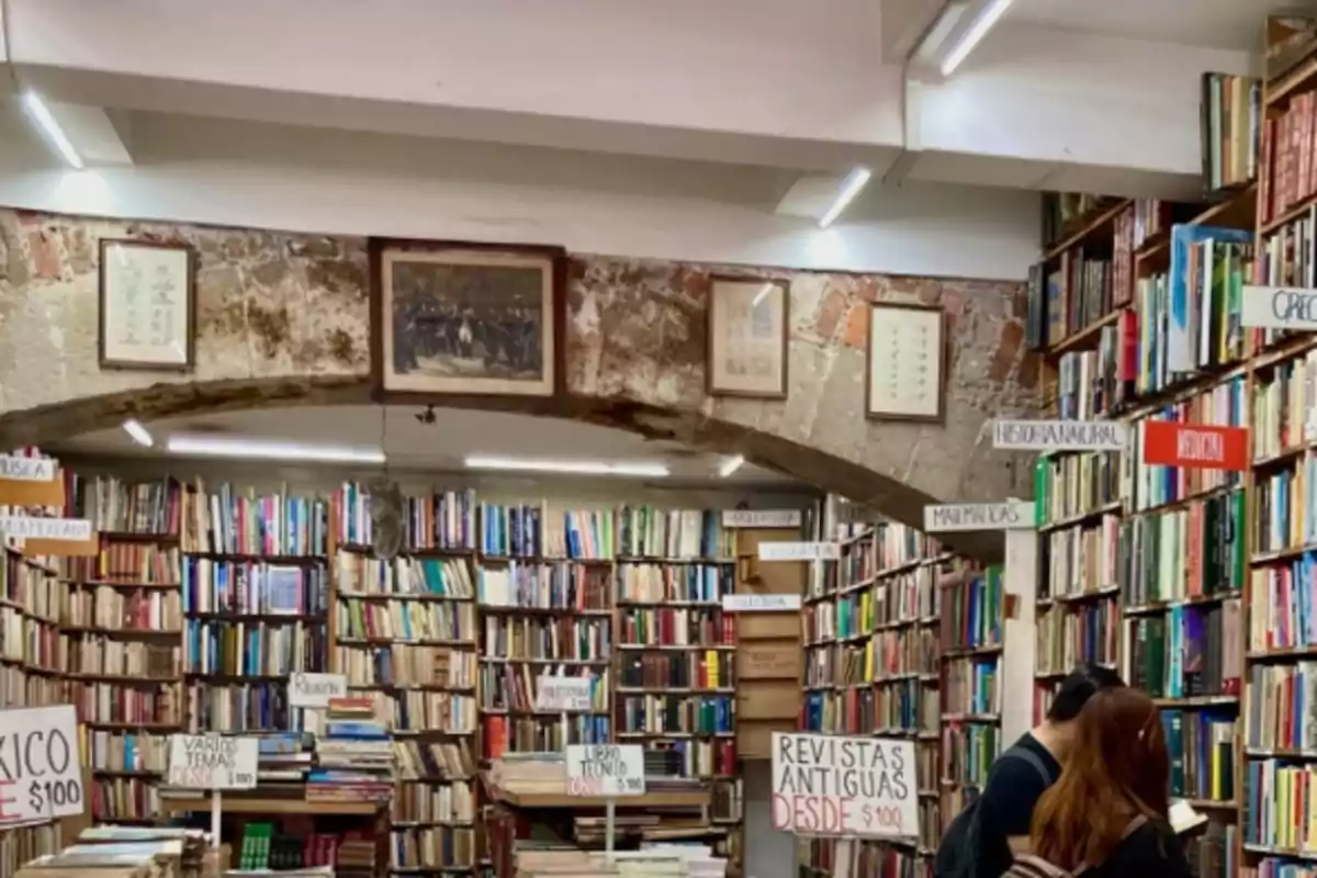 Una librería antigua con estantes llenos de libros y letreros que indican diferentes secciones, con dos personas explorando el lugar.