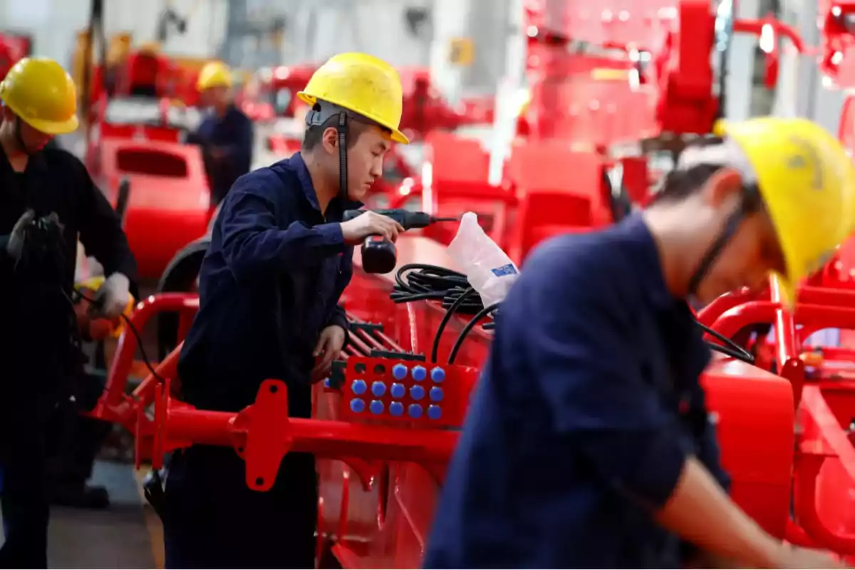 Trabajadores con cascos amarillos ensamblan maquinaria roja en una fábrica.