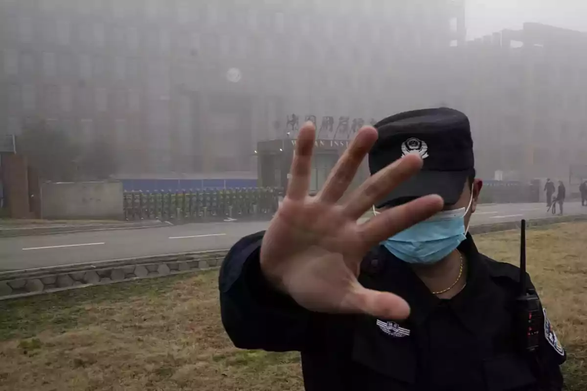 A guard wearing a mask raises his hand in front of the camera on a cloudy day in front of a large building.