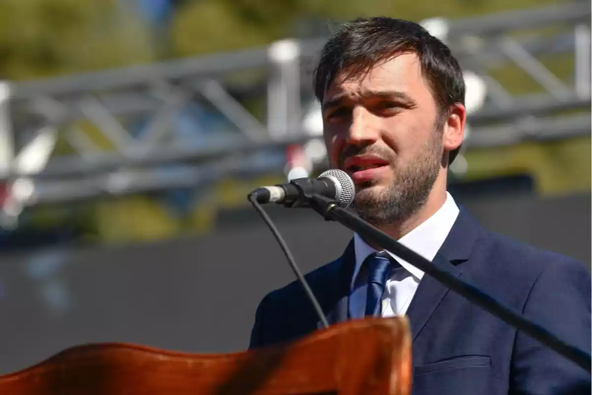 A man in a suit and tie speaks into a microphone during an outdoor event.