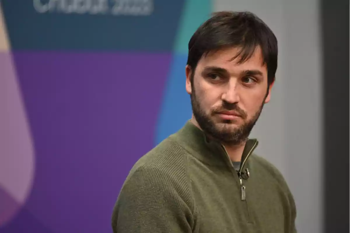 Un hombre con barba y suéter verde mira hacia un lado con expresión seria, fondo con colores morado y azul.