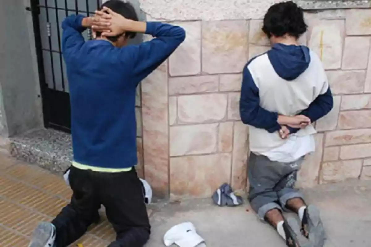 Two people kneeling with their backs against a wall, one with hands on their head and the other with hands behind their back.