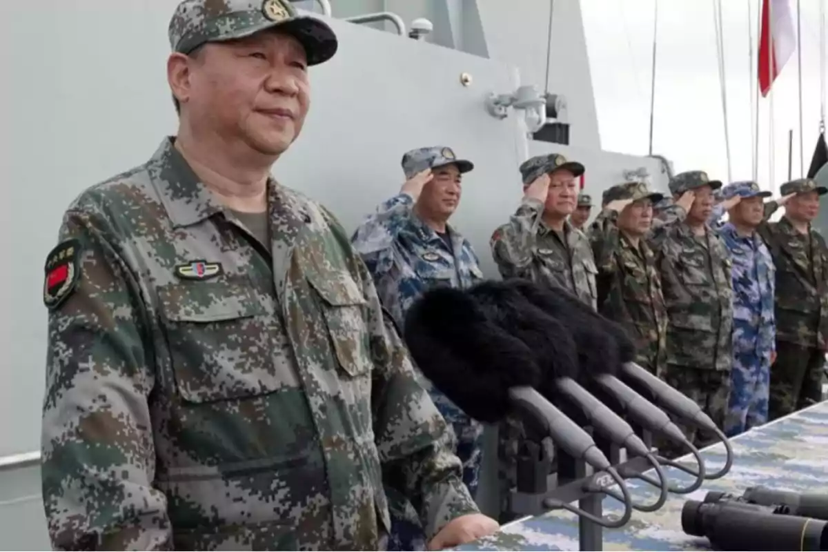 A group of soldiers in camouflage uniforms is standing in a row, some saluting, in front of a set of microphones.