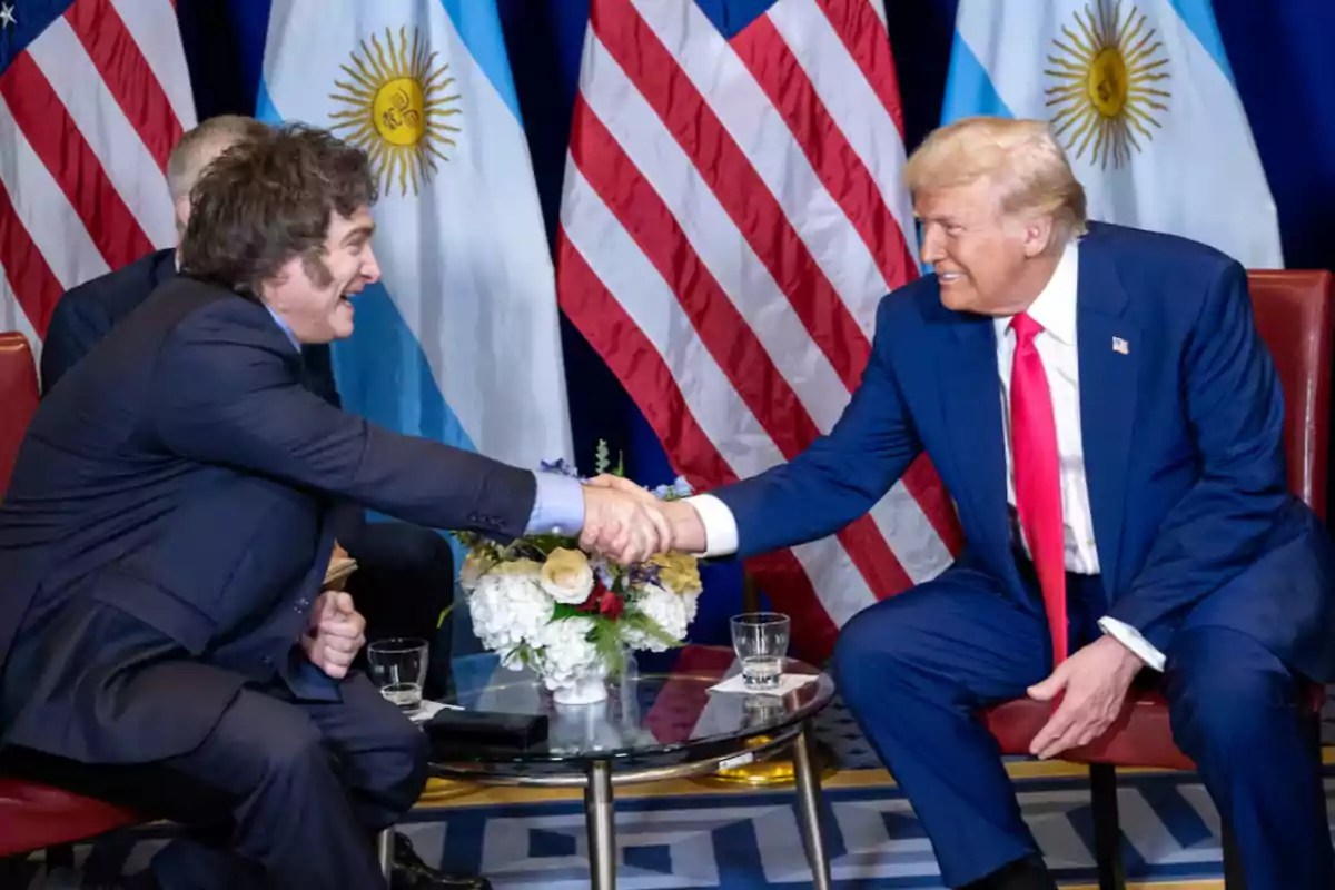 Two people shaking hands at a meeting with the flags of Argentina and the United States in the background.