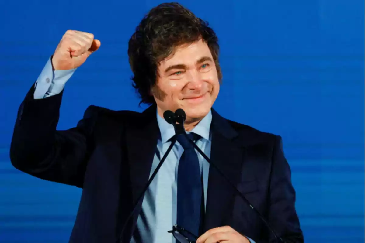 A smiling man in a dark suit and blue tie raises his fist in celebration against a blue background.