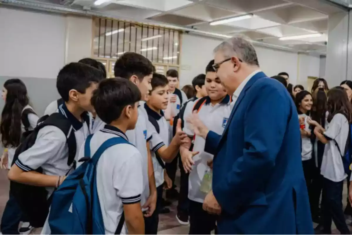 Un grupo de estudiantes con uniformes escolares interactúa con un hombre de traje en un pasillo.