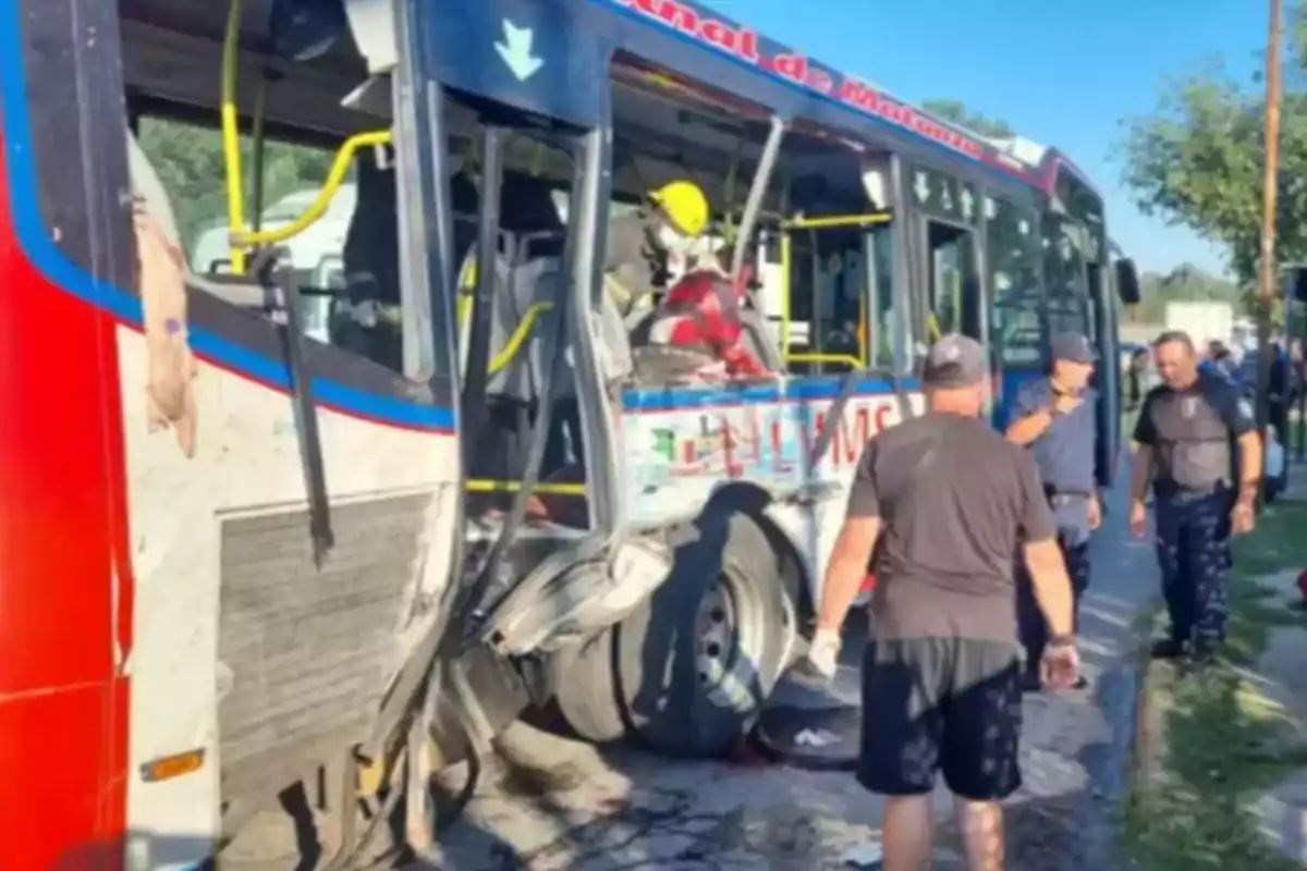 Un autobús con daños significativos en el lateral y varias personas alrededor, incluyendo un bombero con casco amarillo.