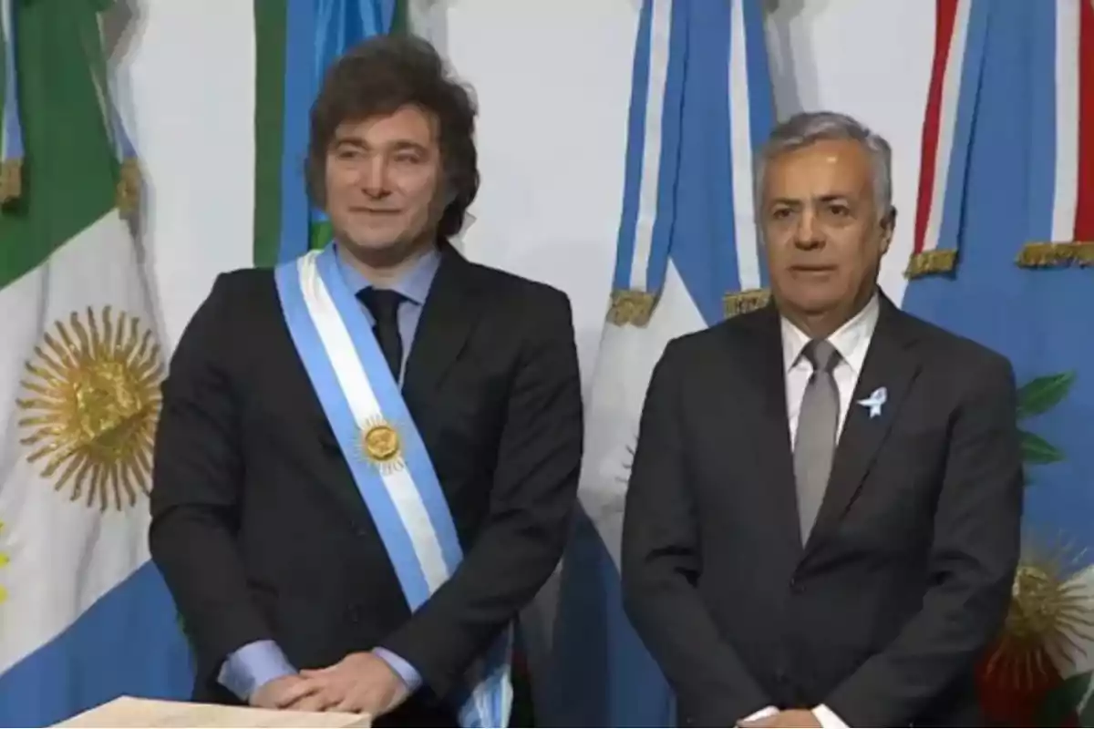 Dos hombres de pie con trajes formales frente a banderas argentinas.