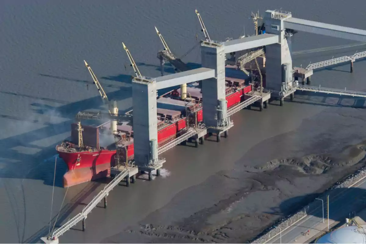 Un barco de carga rojo atracado en un puerto industrial con estructuras de carga y descarga sobre el agua.