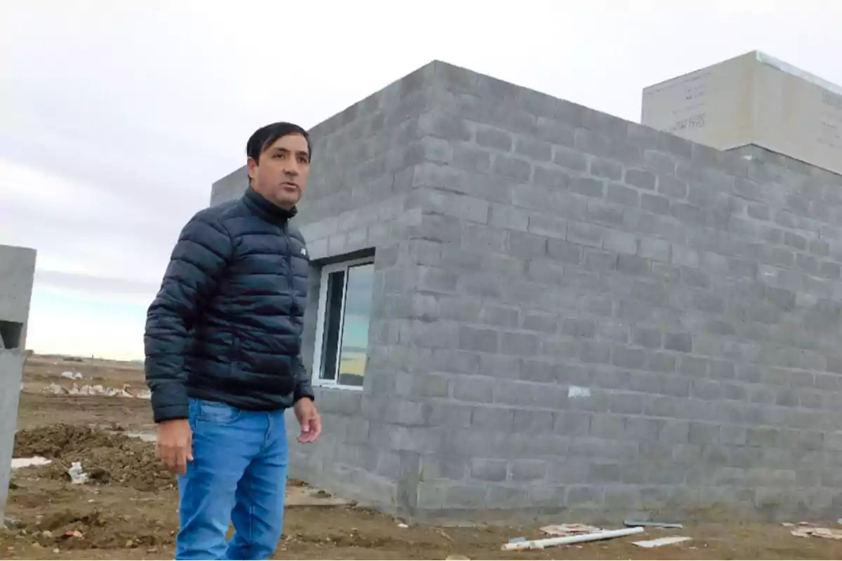 Un hombre con chaqueta negra y jeans está de pie frente a una construcción de bloques de cemento en un área abierta.