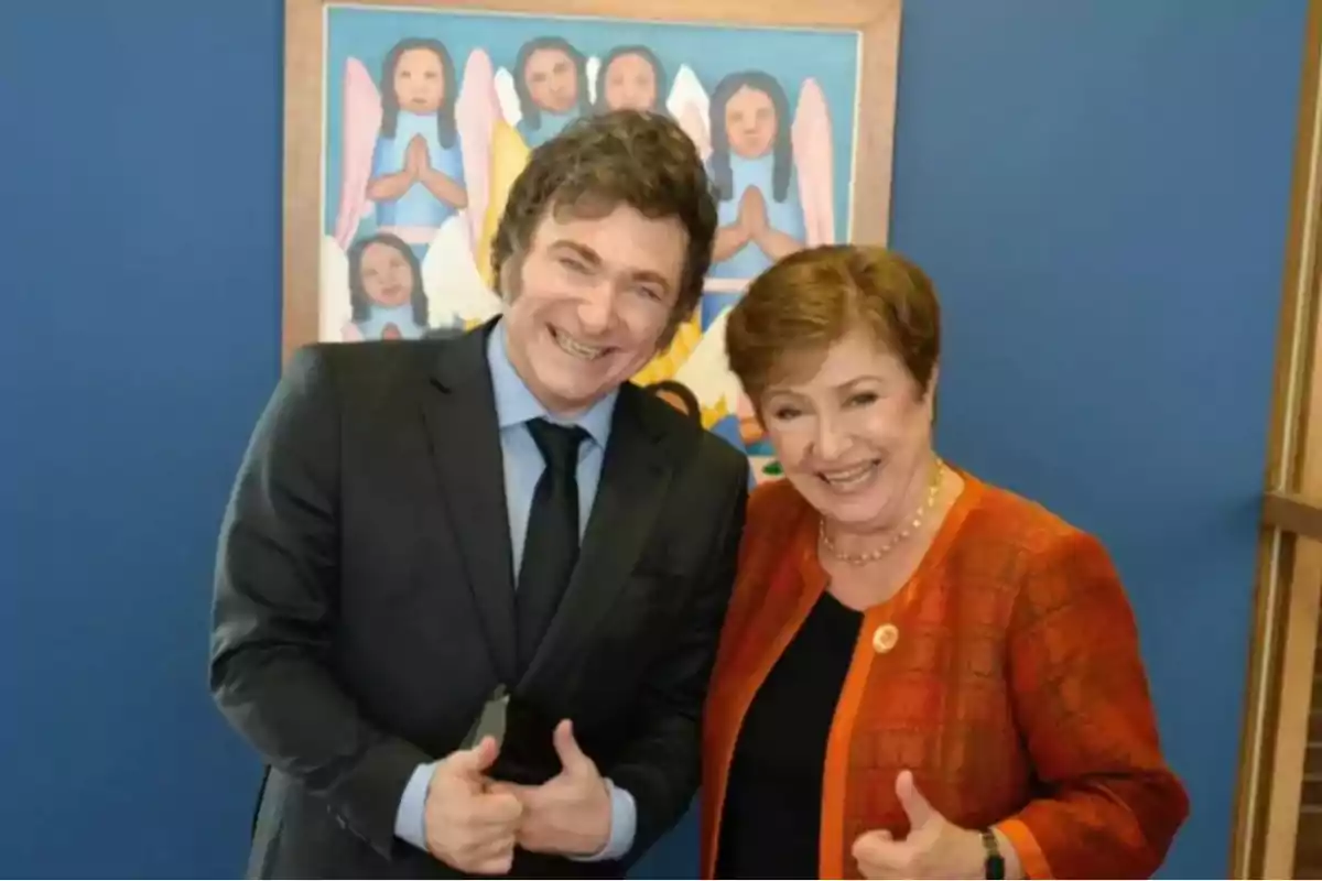 Dos personas sonrientes posan juntas haciendo el gesto de pulgares arriba frente a una pintura colorida de ángeles.