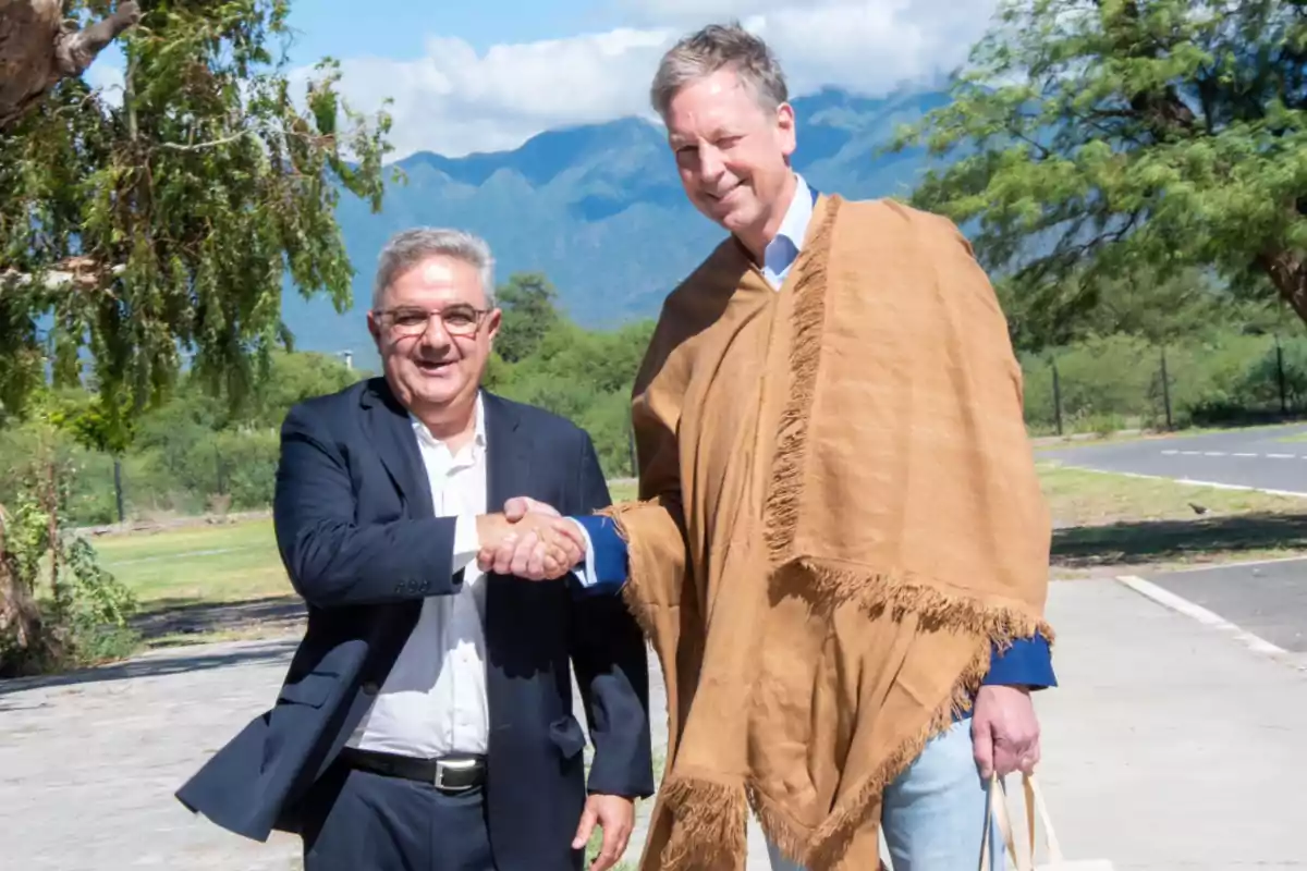 Dos hombres dándose la mano al aire libre con montañas y árboles de fondo.