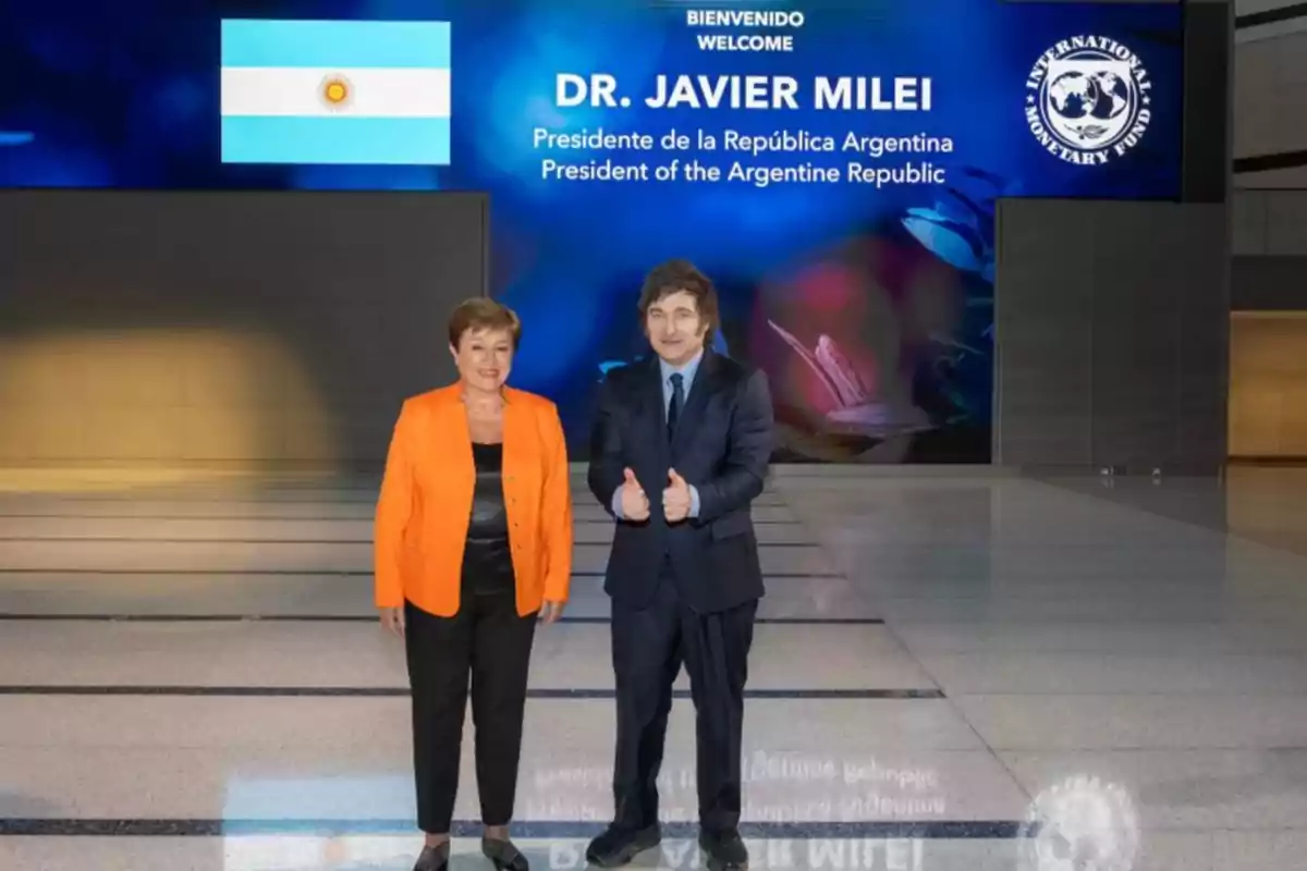 Dos personas posan frente a un cartel de bienvenida que menciona a un presidente de la República Argentina, con la bandera argentina y el logo del Fondo Monetario Internacional.