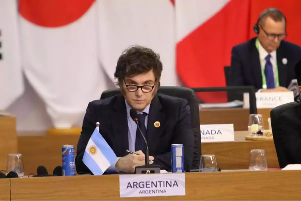 Un hombre con gafas y patillas está sentado en una mesa de conferencia con una bandera de Argentina frente a él y otras personas al fondo.