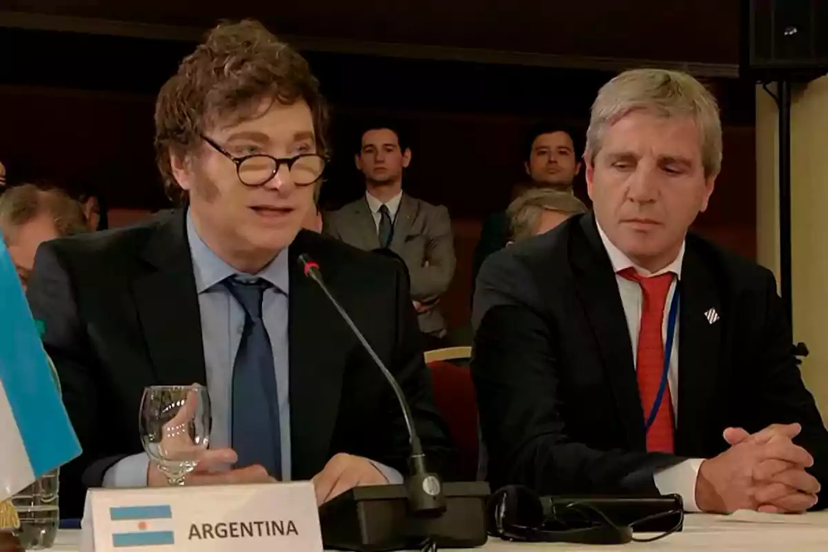 Dos hombres en traje participan en una conferencia, uno de ellos está hablando frente a un micrófono con una bandera de Argentina en la mesa.