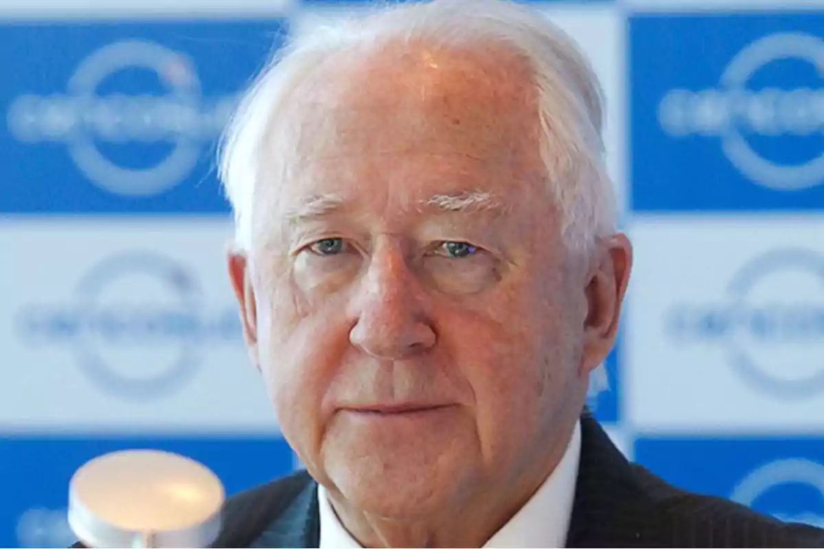 An older man with white hair and a serious expression, in front of a background with blue logos.