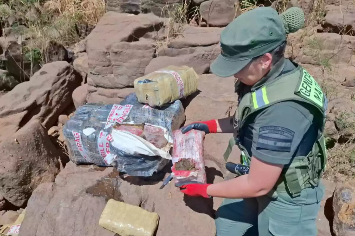 Una persona uniformada inspecciona paquetes envueltos en plástico sobre unas rocas en un entorno al aire libre.